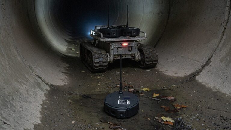 A repeater on the floor of a culvert while a tracked robot drives on into the dark with more repeaters on its deck